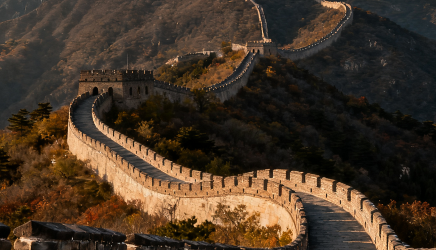 A section of the Great Wall of China winding through mountainous hills, with autumn foliage on the trees and a clear sky above, showcasing the ancient stone structure’s path.